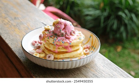 A stack of fluffy pancakes topped with pink ice cream, colorful sprinkles, and cereal rings, served on a white plate. Set against a lush outdoor background, - Powered by Shutterstock