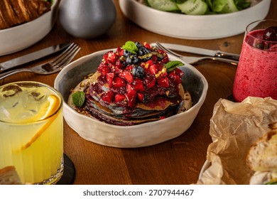 A stack of fluffy pancakes topped with vibrant berry sauce, fresh fruit and mint, surrounded by drinks and breakfast dishes for a cozy brunch setting.