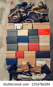 A Stack Of Folded Cardboard Boxes And A Lot Of Variety Of Shoes On The Background Of The Shabby Wall.