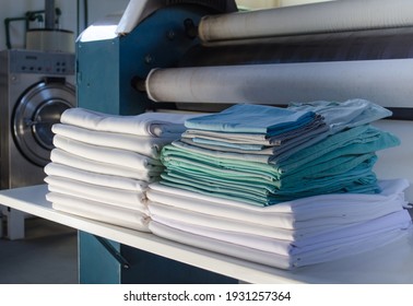 Stack Of Folded Clean Sheets Or Fabrics And Industrial Iron In An Industrial Laundry. Cleaning And Ironing Service For Hotels, Hospitals, Clinics And Companies.