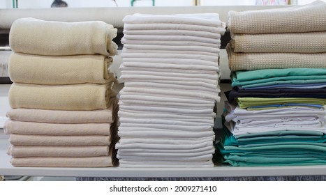 Stack Of Folded Clean Sheets, Surgical Clothes  And Industrial Iron In An Industrial Laundry. Cleaning And Ironing Service For Hospitals And Clinics.