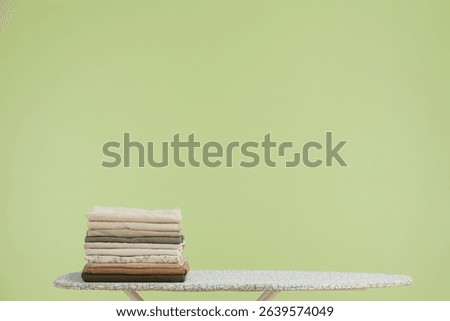 A stack of folded clothes rests on an ironing board against a calm green background.  The muted colors evoke a sense of order and tranquility.