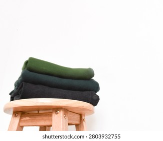 Stack of folded green, dark green, and black clothes on a wooden stool against a minimalist white background wall interior