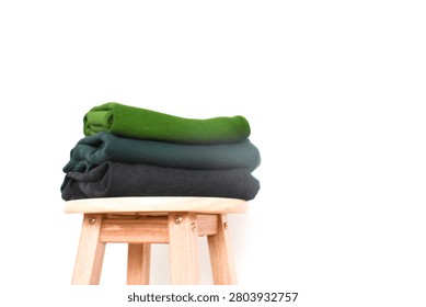 Stack of folded green, dark green, and black clothes on a wooden stool against a minimalist white background wall interior
