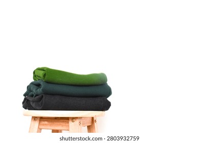 Stack of folded green, dark green, and black clothes on a wooden stool against a minimalist white background wall interior