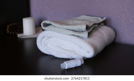 A stack of folded white and light green terry towels, a spool of white thread on a dark wooden table, a candle and a cone on a blurred background.