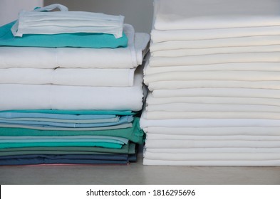 Stack Of Folded White Linens And Green Surgery Clothes In An Industrial Laundry. Clean And Disinfected Clothing Against Covid-19 For Hospitals And Medical Institutions.