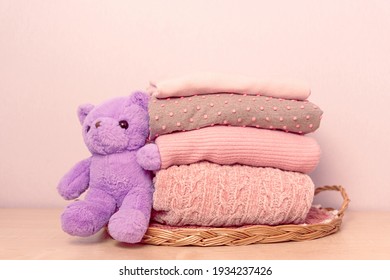 Stack of folded wool knitted sweaters or pullovers in pink pastel colors on table with soft toy Taddy bear. Close up of warm cozy comfortable clothes for autumn winter season. Selective soft focus.