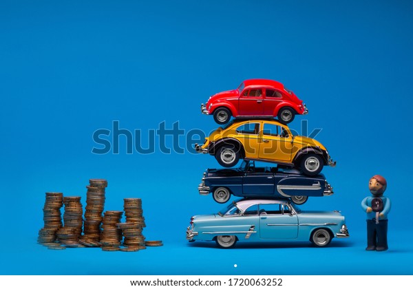 Stack of four car figurines placed in between\
a pile of golden coins and a tiny man made from plasticine, on blue\
background.