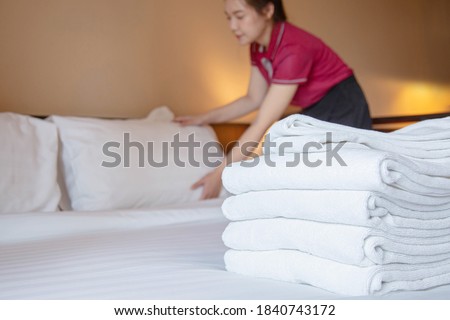A stack of fresh towels is on the twin bedroom while hotel maid is setting pillows in the background. Focus on a stack of towels.