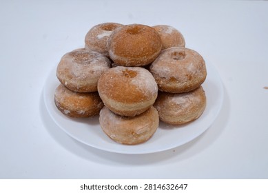A stack of freshly fried glazed donuts dusted with powdered sugar, arranged on a white round plate against a clean white background. The homemade doughnuts show a golden-brown fried exterior with a light sugar coating, piled in a pyramid shape with visible center holes.