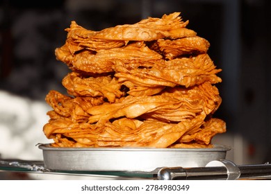 Stack of fried, golden-brown, crispy snack pieces. Food sits on metal plate. Warm lighting highlights texture. Appears freshly cooked and ready to eat