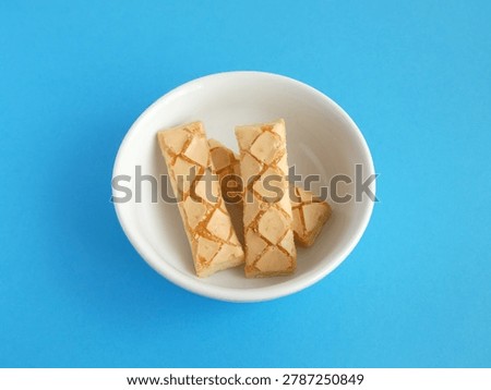 Stack of glazed criss-cross pattern puff pastry cookies in white bowl. Top view of several crispy rectangular puff pastry biscuits with sweet glazed criss-cross patterns piled together in a white bowl on a blue background.