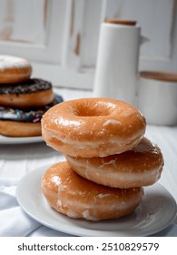 stack of glazed donuts placed on a white plate. In the background, there is another plate with a variety of donuts, including one with chocolate sprinkles. The scene is set in a light, airy kitchen.
