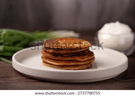 stack of golden-brown pancakes with crispy edges on a simple white plate, photographed on a dark wooden tabletop with blurred scallions and a glass bowl of sour cream in warm, rustic bokeh lighting