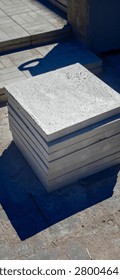 Stack of gray concrete slabs arranged neatly on a construction site with a textured surface and shadow cast by nearby structures in bright sunlight