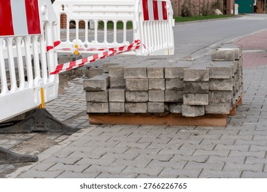 Stack of grey concrete paving blocks on wooden pallet beside white safety barriers with red tape at urban road repair site. Concept of street reconstruction, paving materials supply . 