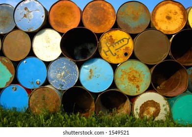A stack of grungy old oil drums at a music festival, ready to be set out as rubbish bins. One of them is for used syringes. Blue sky above and behind, green grass below.