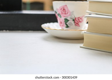 Stack of hardback books and a cup of tea in a vintage china cup and saucer on a windowsill. Selective focus.