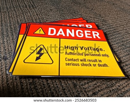 Stack of high voltage warning signs on a gray carpeted floor