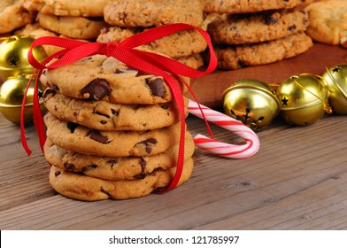 A Stack Of Holiday Chocolate Chip Cookies Tied With A Red Ribbon In Front Of Jingle Bells , Candy Cane, And A Platter Of Assorted Baked Treats. Horizontal Format With Shallow Depth Of Field.