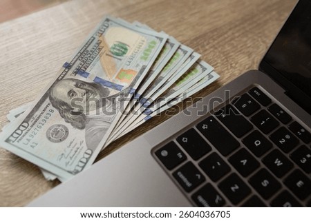 A stack of hundred dollar bills placed on a wooden surface next to a laptop keyboard. Concept of online earnings, digital finance and remote work. High-quality photo. High quality photo