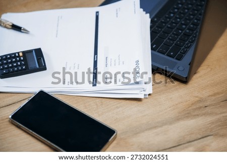 Stack of invoice documents on a wooden desk with a calculator, a pen, a phone, and a laptop. Business accounting concept for financial management.