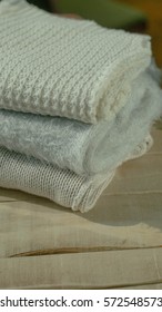 Stack of knitting sweaters in white and grey tones on the light wooden table. Warm clothes. Knitted background.