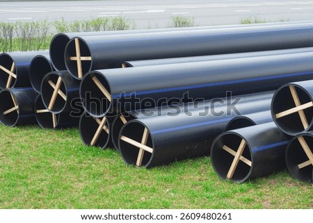 Stack of large black pipes for water mains with blue stripes, rests on a grassy field