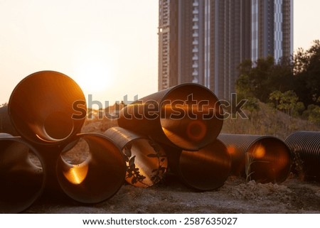 a stack of large pipes arranged at a construction site, with the setting sun casting light on them. Tall buildings can be seen in the background, and some plants are growing around the pipes.