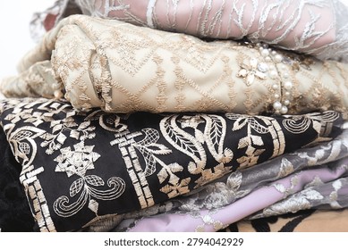Stack of luxury fabrics in soft tones and intricate patterns. Beige, black, and silver textures showcase elegance and craftsmanship. Close-up highlights embroidery, lace, and delicate detailing.