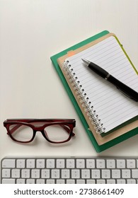 A Stack Of Notebooks With A Black Pen And Reading Glasses And A Silver Computer Keyboard On A Minimalist White Desk