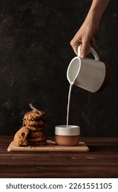 Stack of oatmeal cookies and a cup of milk. Milk is pouring into a cup and fresh home made cookies on a rustic wooden table on a dark background. - Shutterstock ID 2261551105