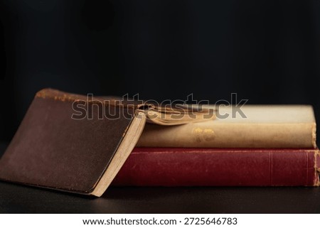 A stack of old books with a brown leather-bound book on top, resting on a dark surface against a black background.