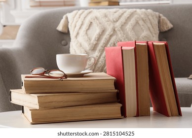 Stack of old books with reading glasses and a cup of coffee on a white table in a cozy living room, Several red-covered books stand upright next to the stack