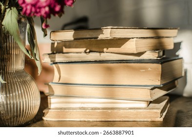 A stack of old books. To the side are wilted pink flowers in a glass vase.