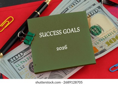 A stack of one hundred dollar bills sits beside a pen, and colorful paperclips. A green notepad proclaims SUCCESS GOALS for 2026 on a red surface.