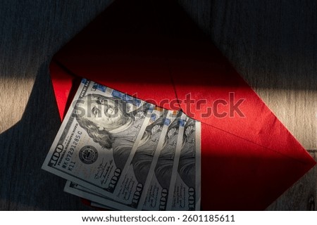 Stack of one hundred dollar bills inside red envelope on wooden surface with dramatic light and shadows. Concept of financial transactions, gift money, bribery, wealth, or business cash flow . 