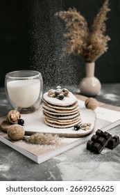 Stack of pancakes with berries and walnuts, dusted with powdered sugar, next to a glass of milk and chocolate pieces on a rustic table.