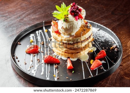 Stack of pancakes with ice cream, mint, and strawberries on a dark plate, drizzled with syrup