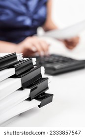 A stack of papers with Binder Clips against the background of an office worker counting on a calculator