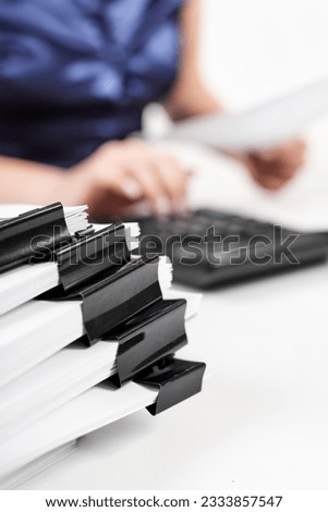 A stack of papers with Binder Clips against the background of an office worker counting on a calculator
