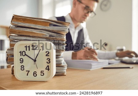 Stack of papers, table clock and businessman engrossed in work, symbolize the overtime and value of time. Image showing dedication, hard work, business environment and importance of time management.