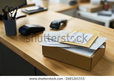 Stack of parcels and envelopes resting on wooden office desk, including cardboard box and various documents, background showing payment terminal and office supplies in workspace