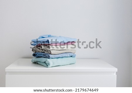 Stack of perfectly folded clothing items. Pile of different cold color shirts, sweaters and pants on the table, white wall background. Close up, copy space, studio shot.