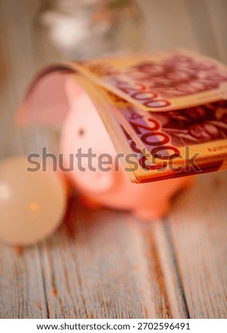 A stack of pink Ghana Cedi (200 GHS) banknotes is placed on top of  a blurry pink piggy bank on a wooden surface with LED bulb, emphasizing savings and wealth accumulation.