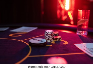 A Stack Of Poker Chips In A Casino On A Table With A Dealer's Mark Nearby, A Cozy, Dark Atmosphere, A Glass Of Water With Lemon Nearby, Red Light On The Background. Casino Chips On Gambling Table. 