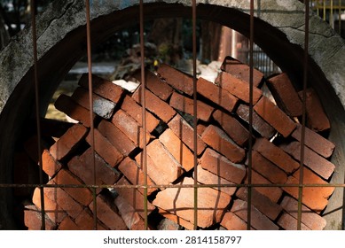 Stack of red bricks inside concrete pipe viewed through metal fence illuminated by dramatic warm sunlight, highlighting their rough texture and varied surface imperfections.