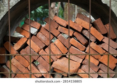Stack of red bricks inside concrete pipe viewed through metal fence illuminated by dramatic warm sunlight, highlighting their rough texture and varied surface imperfections.