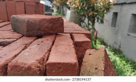 Stack of red bricks outdoors with a single brick on top, near a white wall and greenery